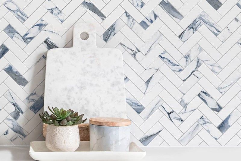 A cutting board and a potted plant are on a shelf in front of a herringbone tile wall.