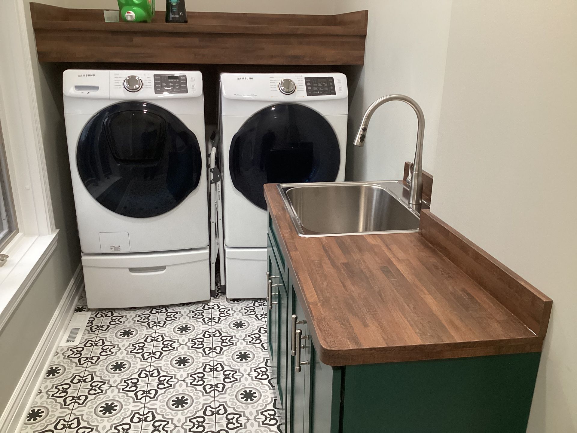Laundry room with washer, dryer, sink, and green cabinet with a wood countertop. Black and white patterned floor.
