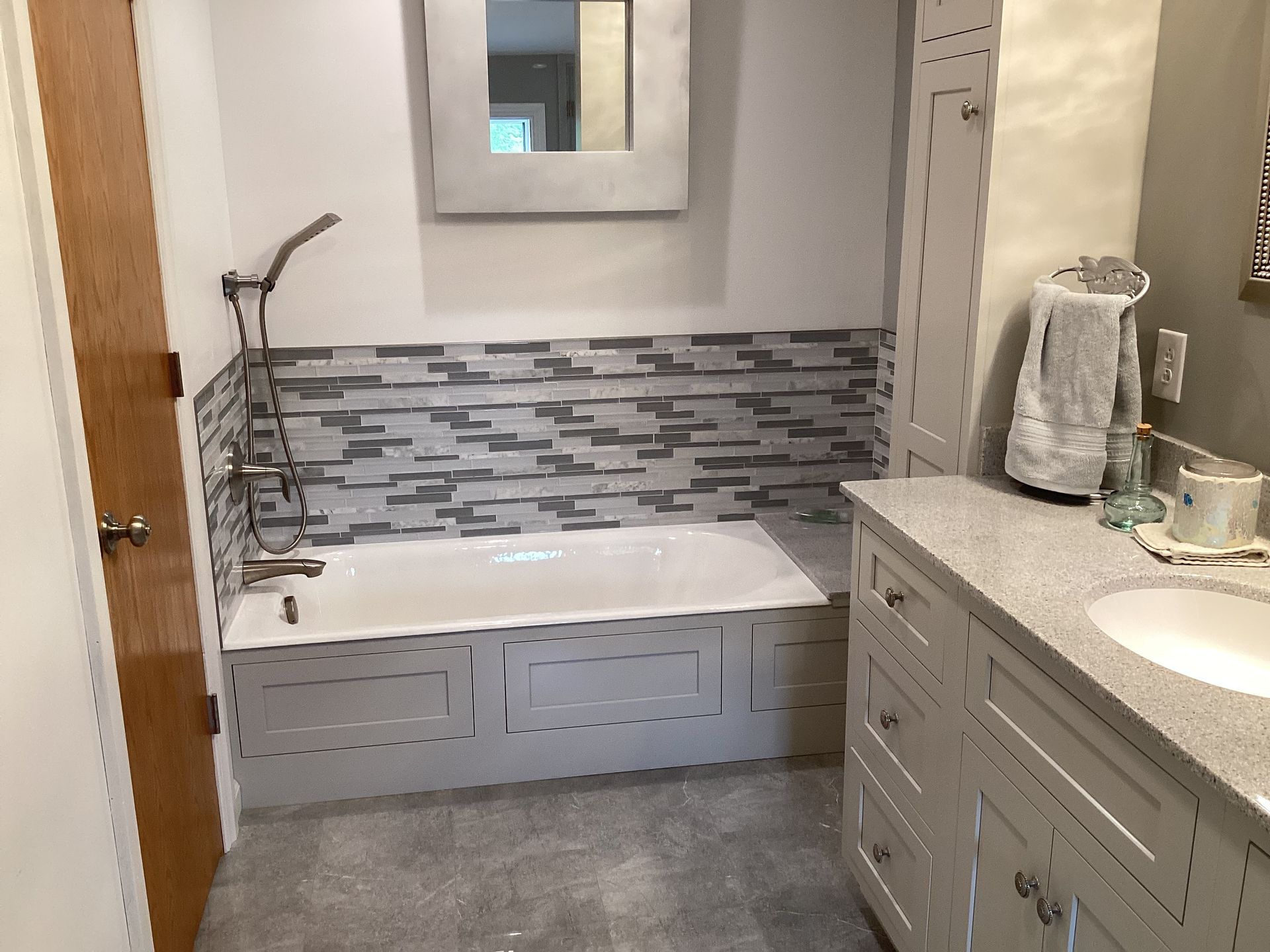 Bathroom with gray cabinets, tub, and mosaic tile backsplash. A towel hangs near the sink.