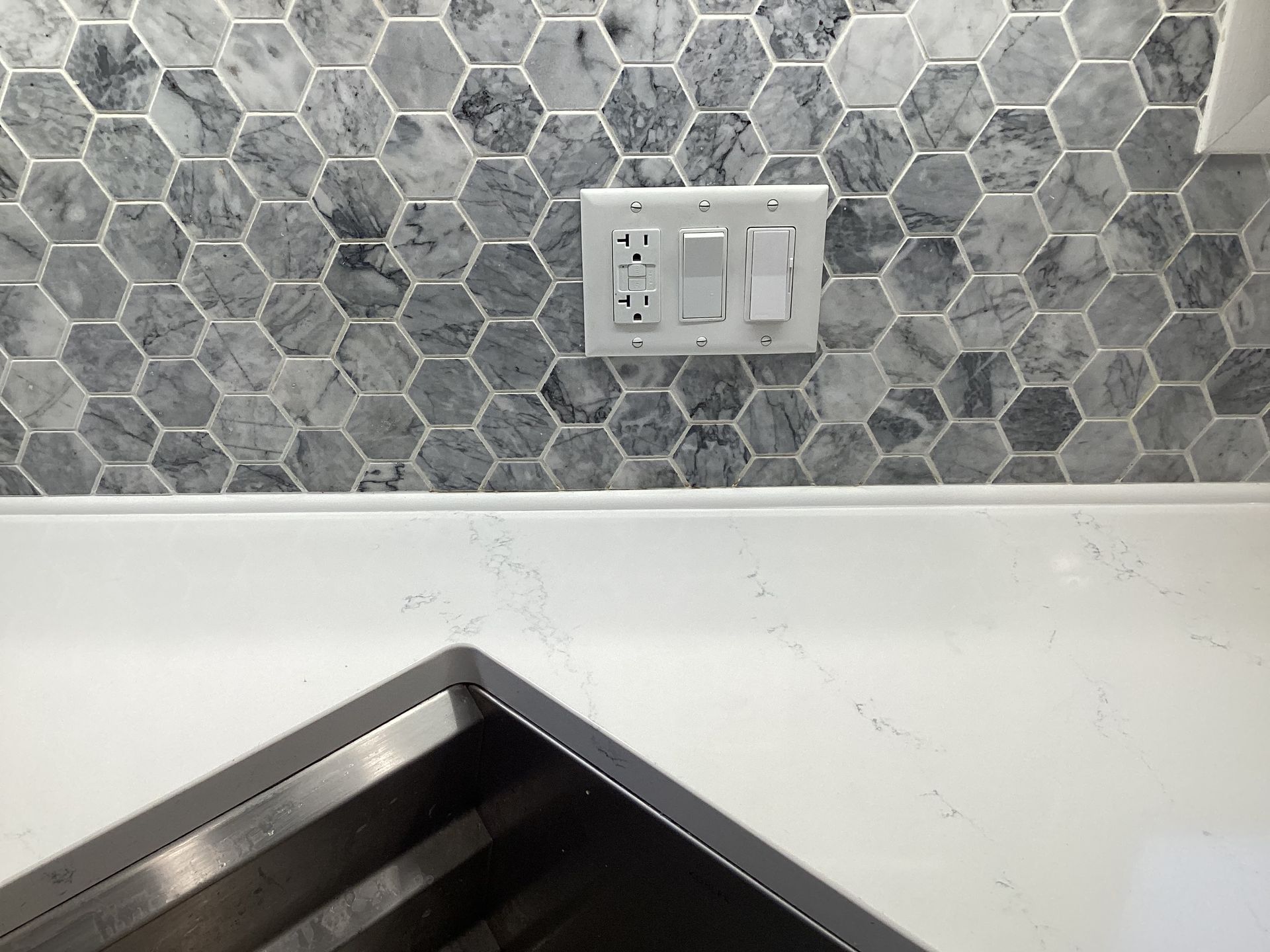 Close-up of a white countertop with a stainless steel sink, a backsplash of gray hexagon tiles, and electrical outlets and a switch.