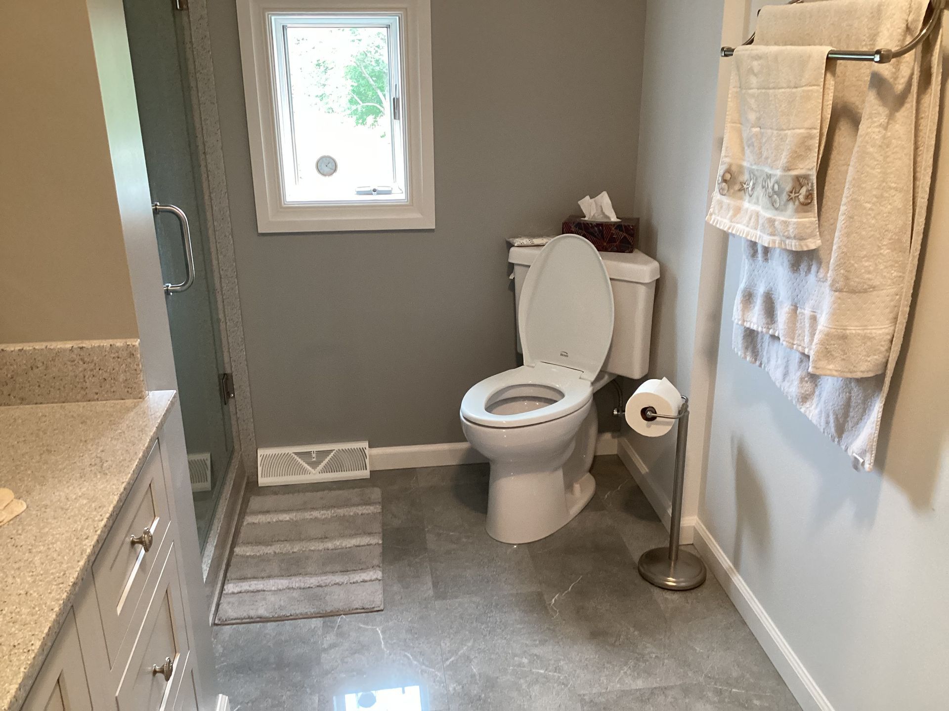 Bathroom with gray walls, toilet, shower, vanity, window, and towels.