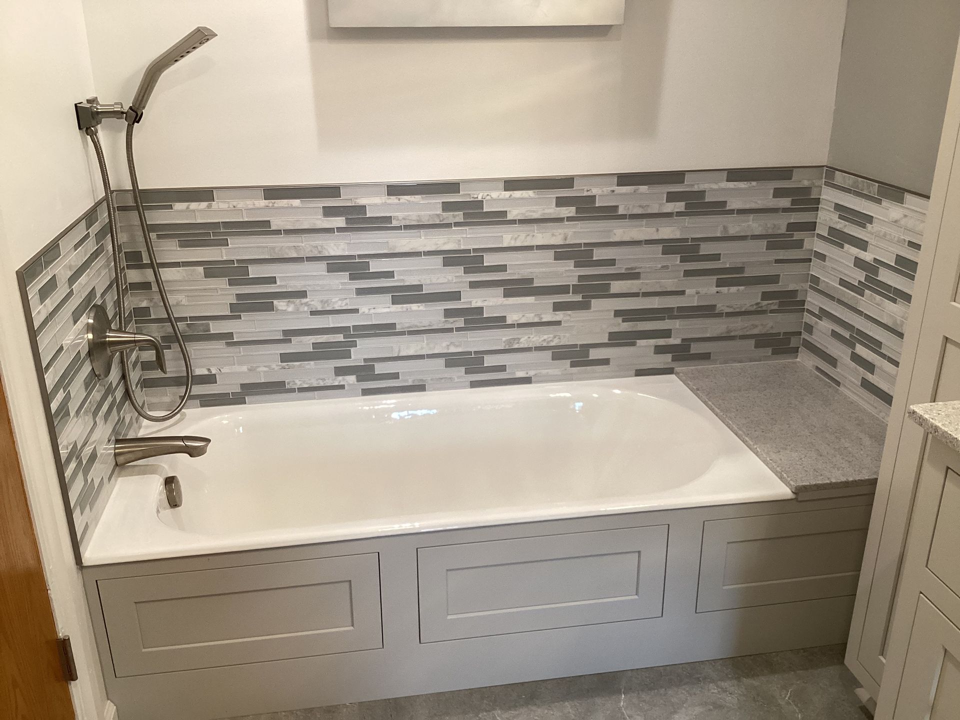 Bathroom with white tub, grey tile backsplash, and light grey paneling.