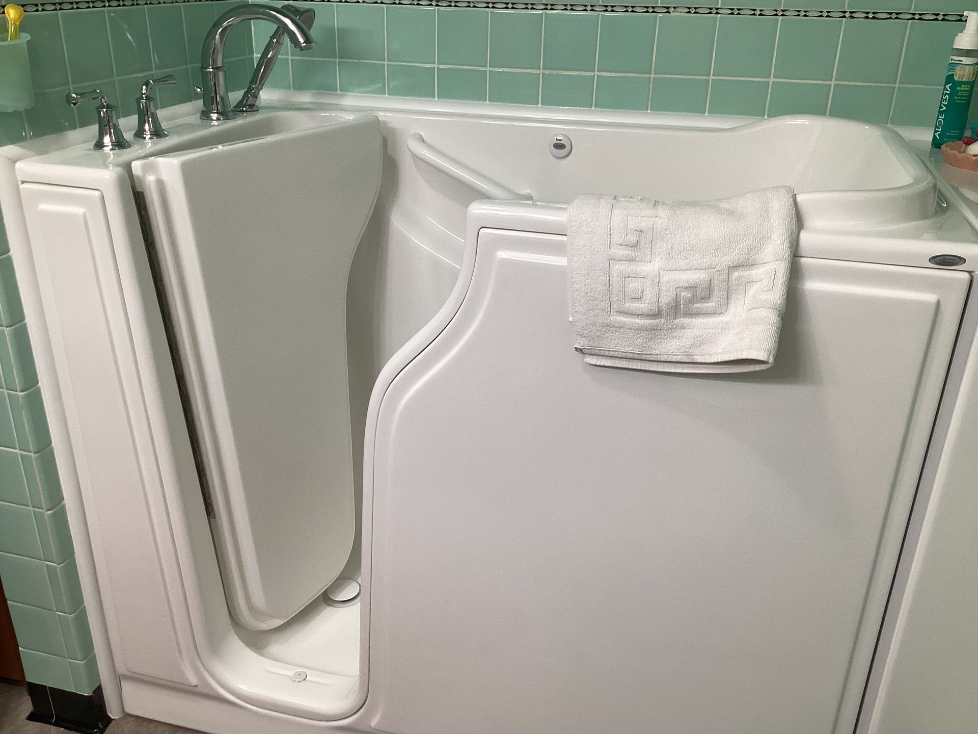 White walk-in bathtub with open door. A white towel hangs on the closed door. The tub sits by a teal tiled wall.
