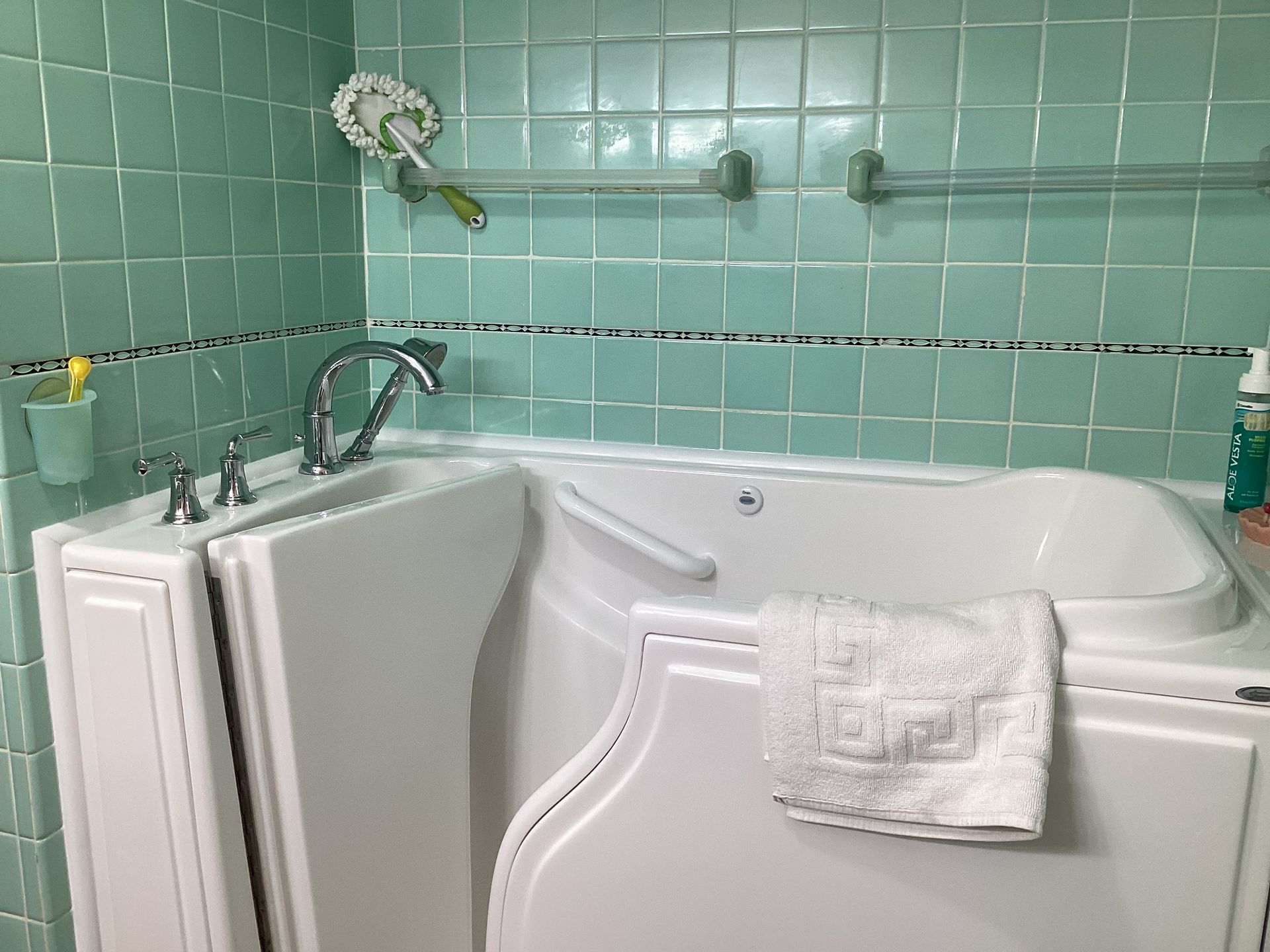 Walk-in bathtub in a tiled bathroom with a towel, showerhead, and cleaning supplies.