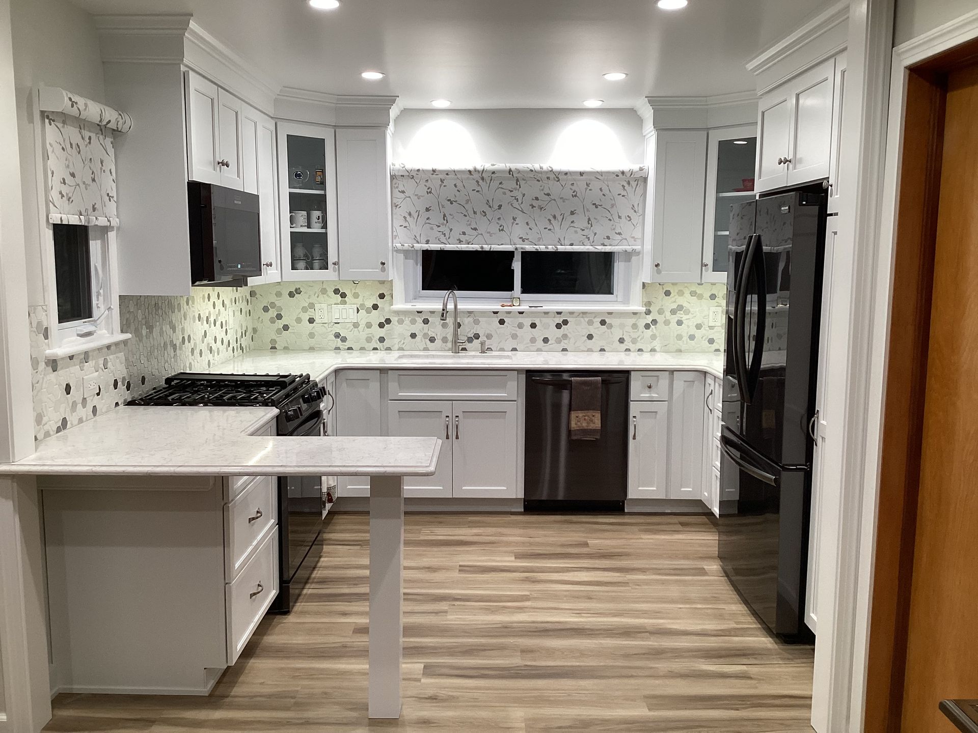 White kitchen with light wood floor, white cabinets, black appliances, speckled backsplash and window shades.