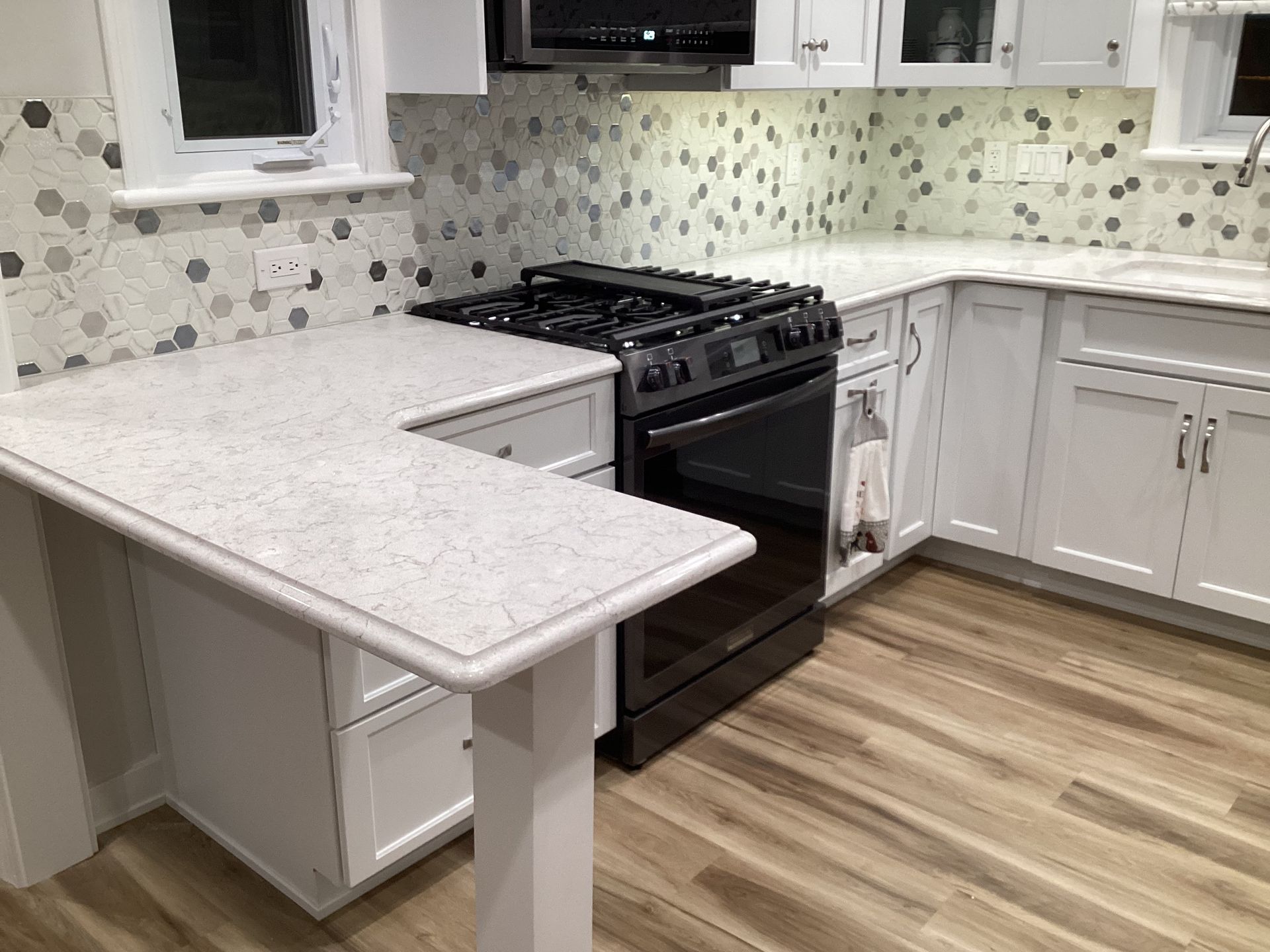 White kitchen with white countertops and cabinets, black stove, gray and white tile backsplash, and light wood floors.