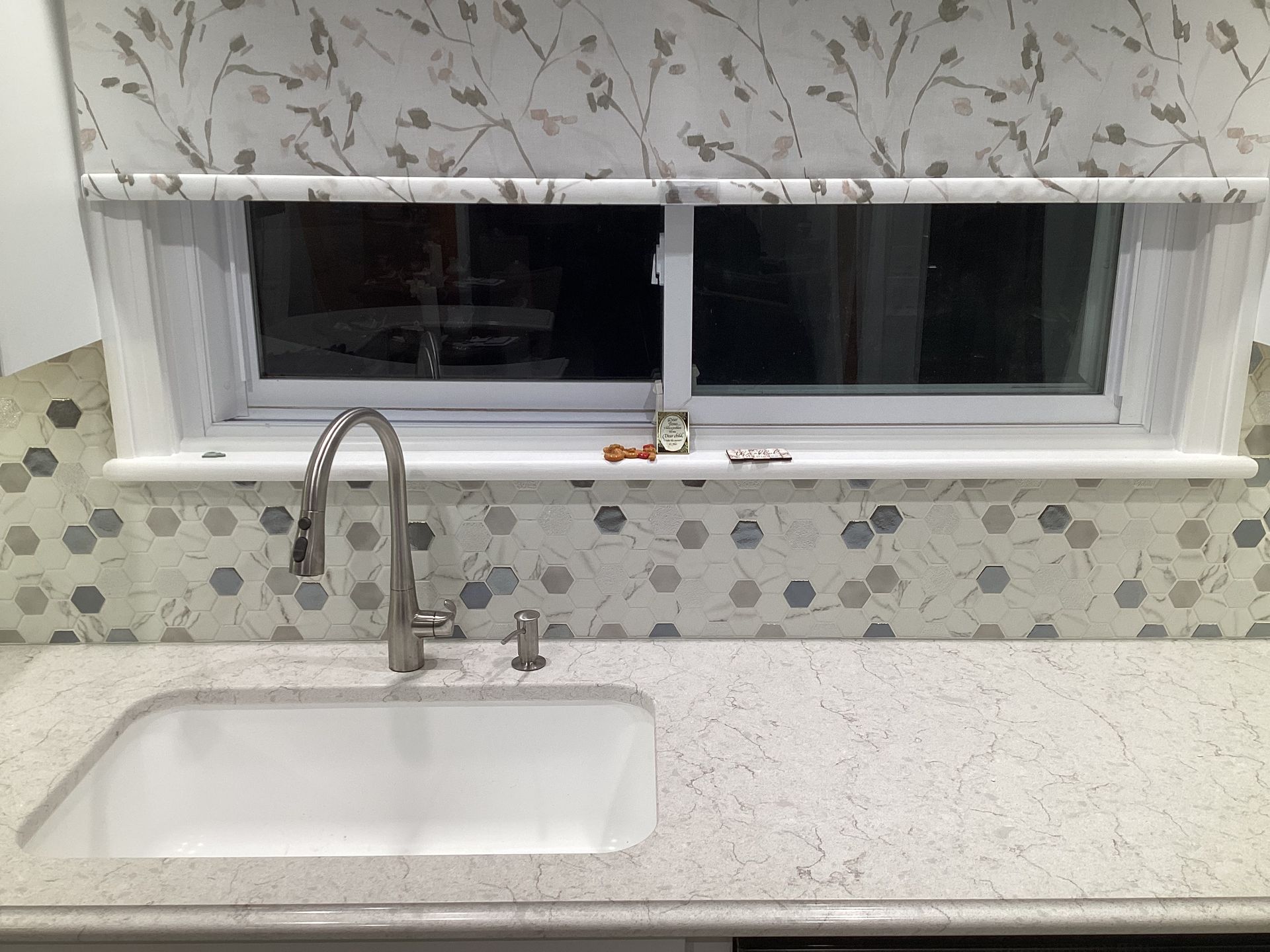 Kitchen sink with faucet, white counter, and backsplash tile. Window with floral print shade.