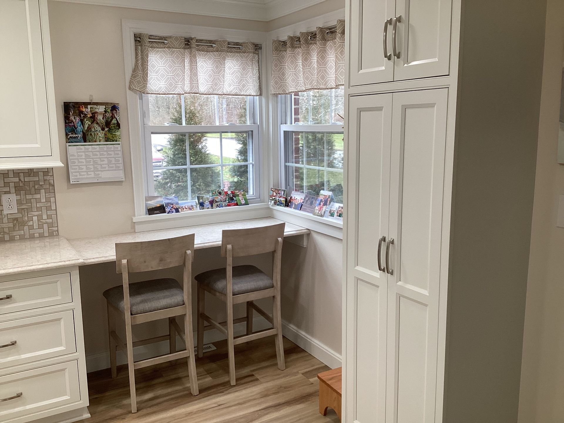 Kitchen nook with countertop desk, two stools, and window with patterned curtains. Tall white storage cabinet.