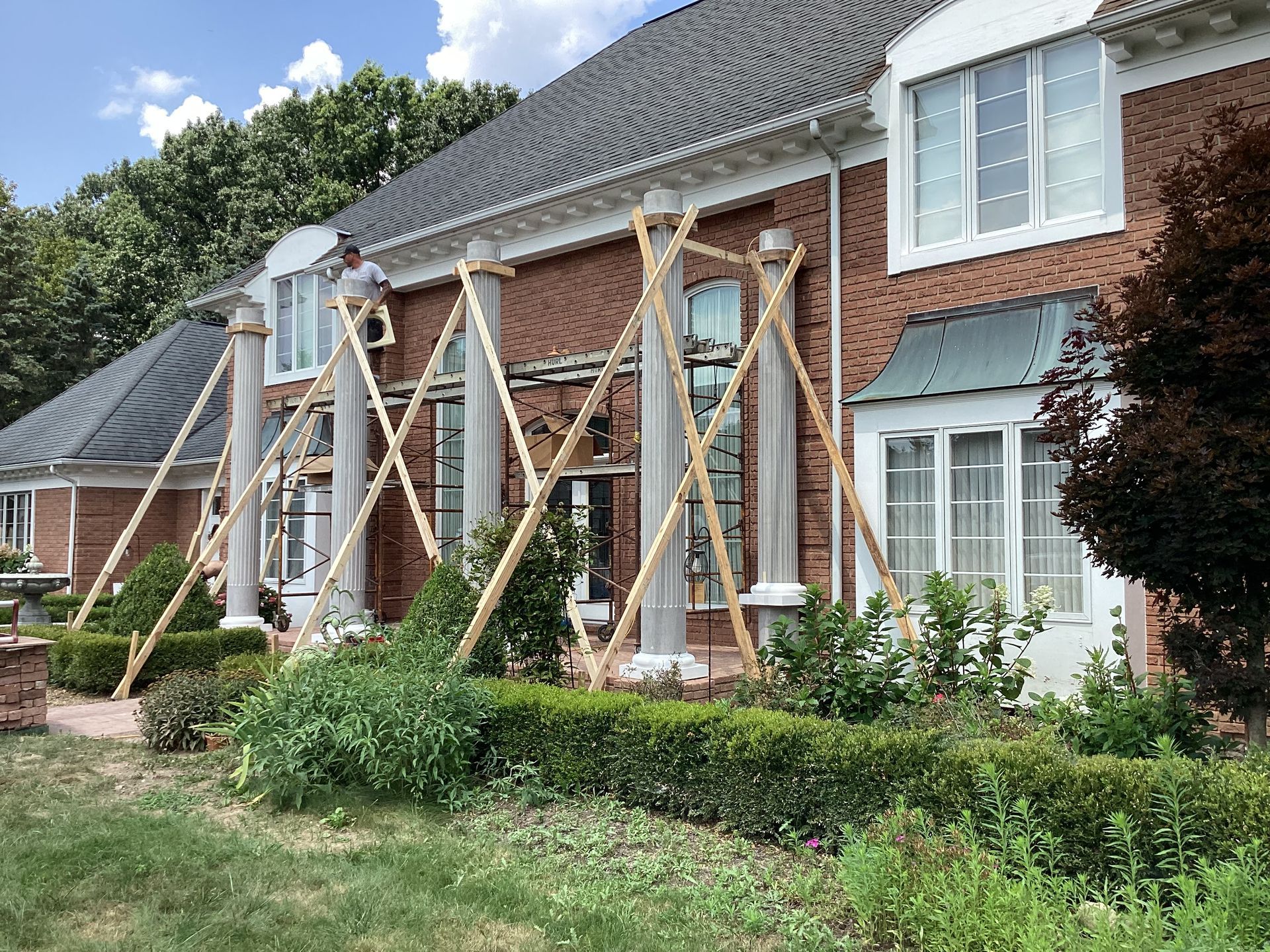 Man on scaffolding working on a brick house with columns, supported by wooden bracing. Green bushes in front.