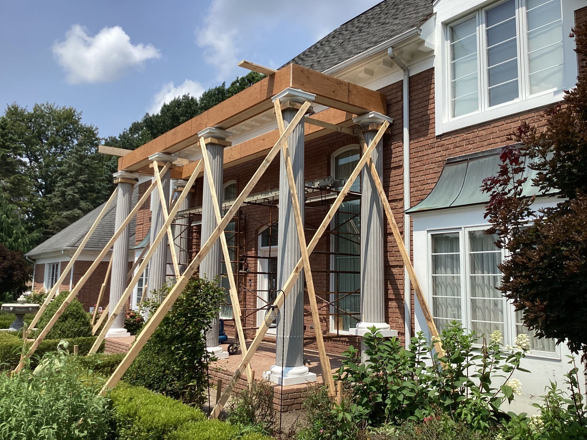 House porch under construction, supported by wooden beams, with brick exterior and white columns.