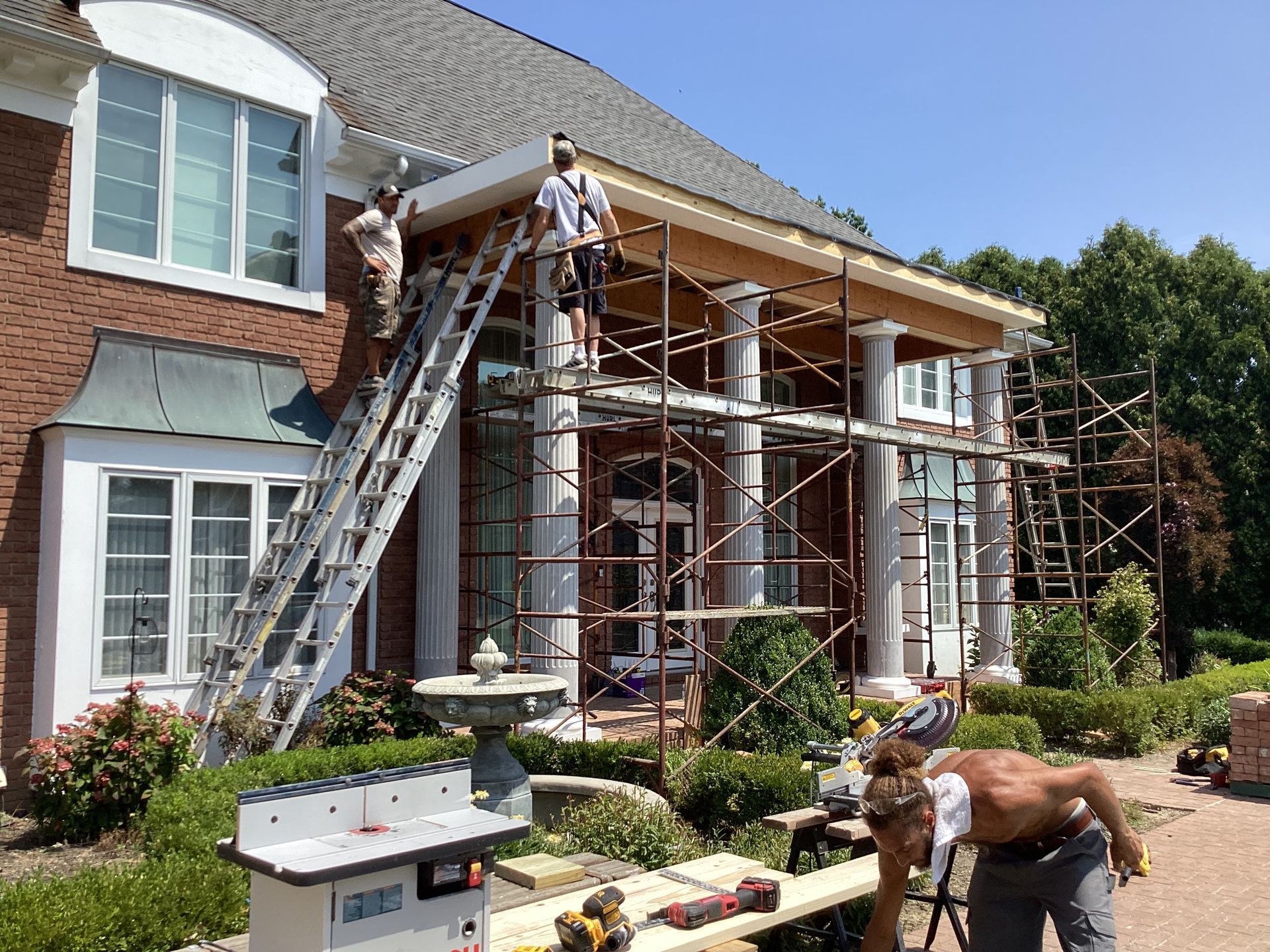 Construction workers building a porch on a brick house. One on a ladder, two on scaffolding. Sunny day.