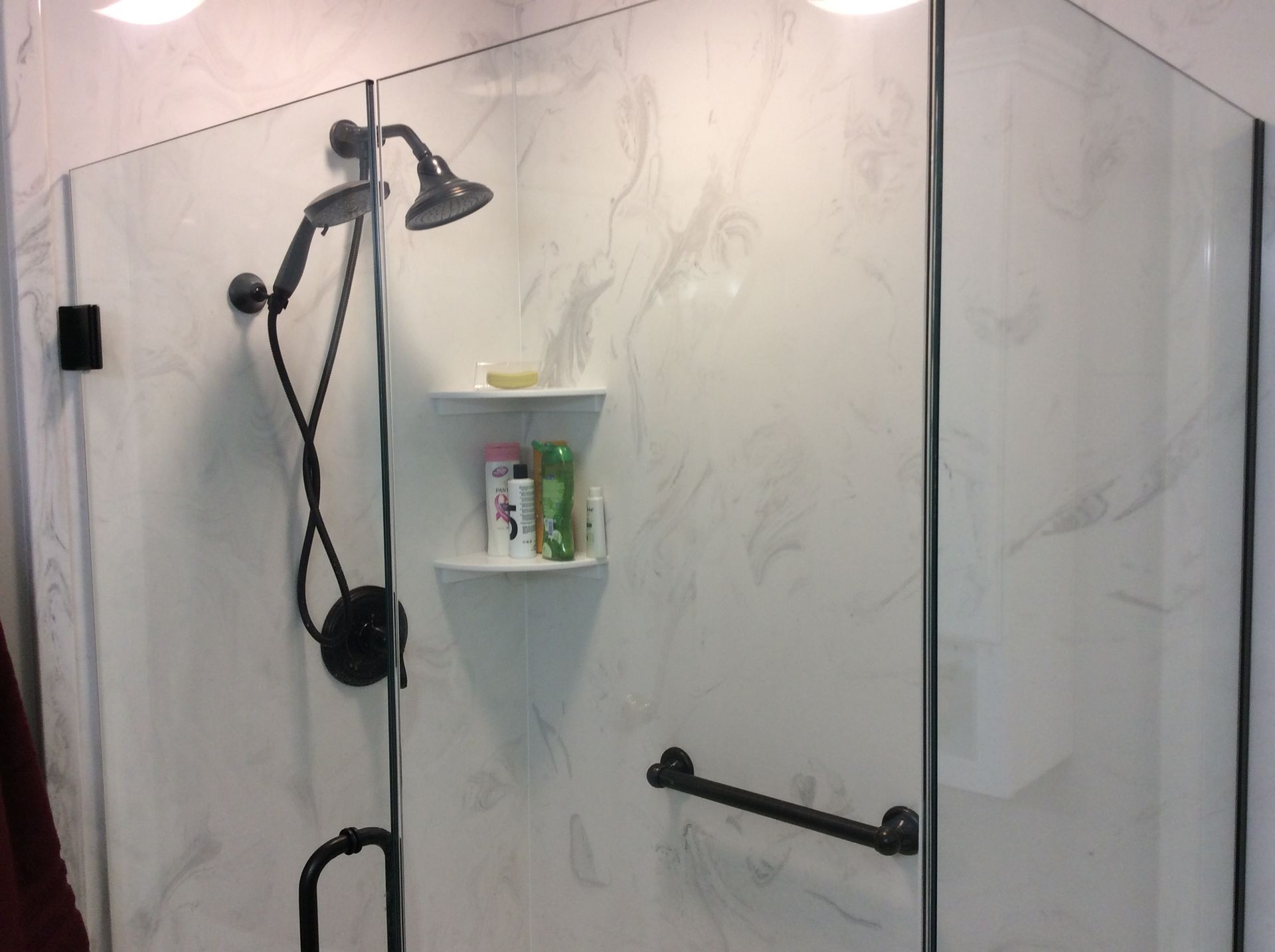 Shower with glass doors, black fixtures, marble-look walls, and a built-in shelf with toiletries.