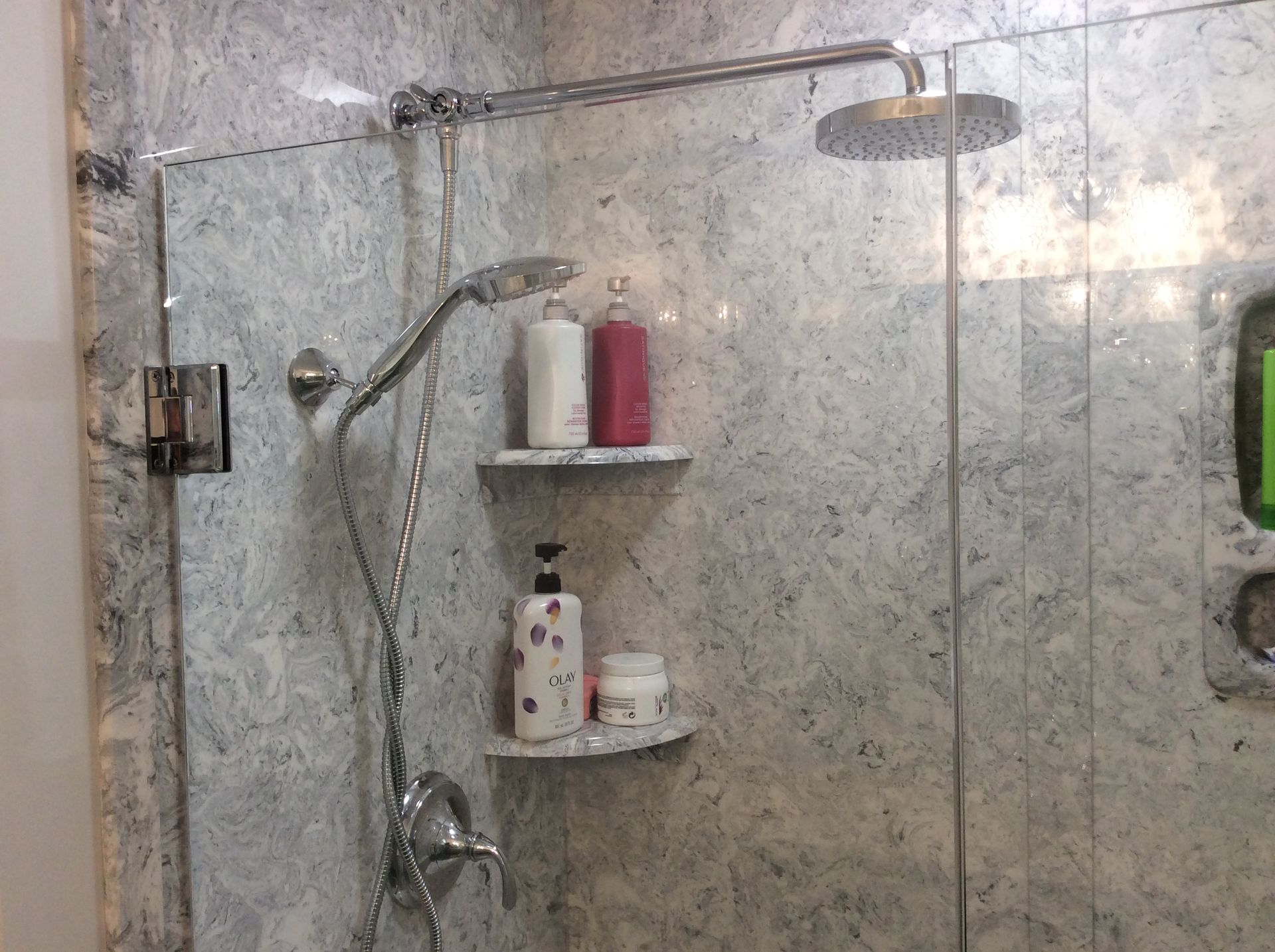 Shower with marble-like wall, glass door, and rain shower head. Features shelves with shampoo bottles.