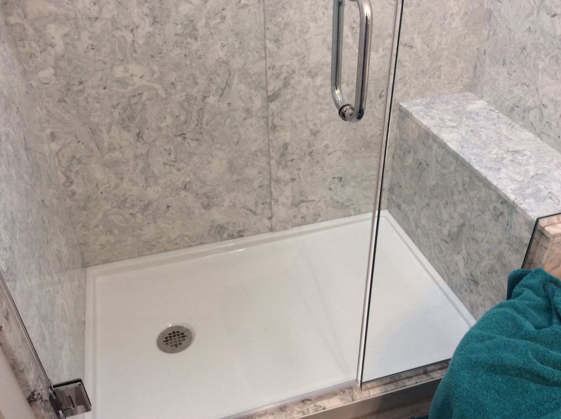 Shower stall with gray marble walls, white floor, glass door, and built-in bench. A teal towel is visible.