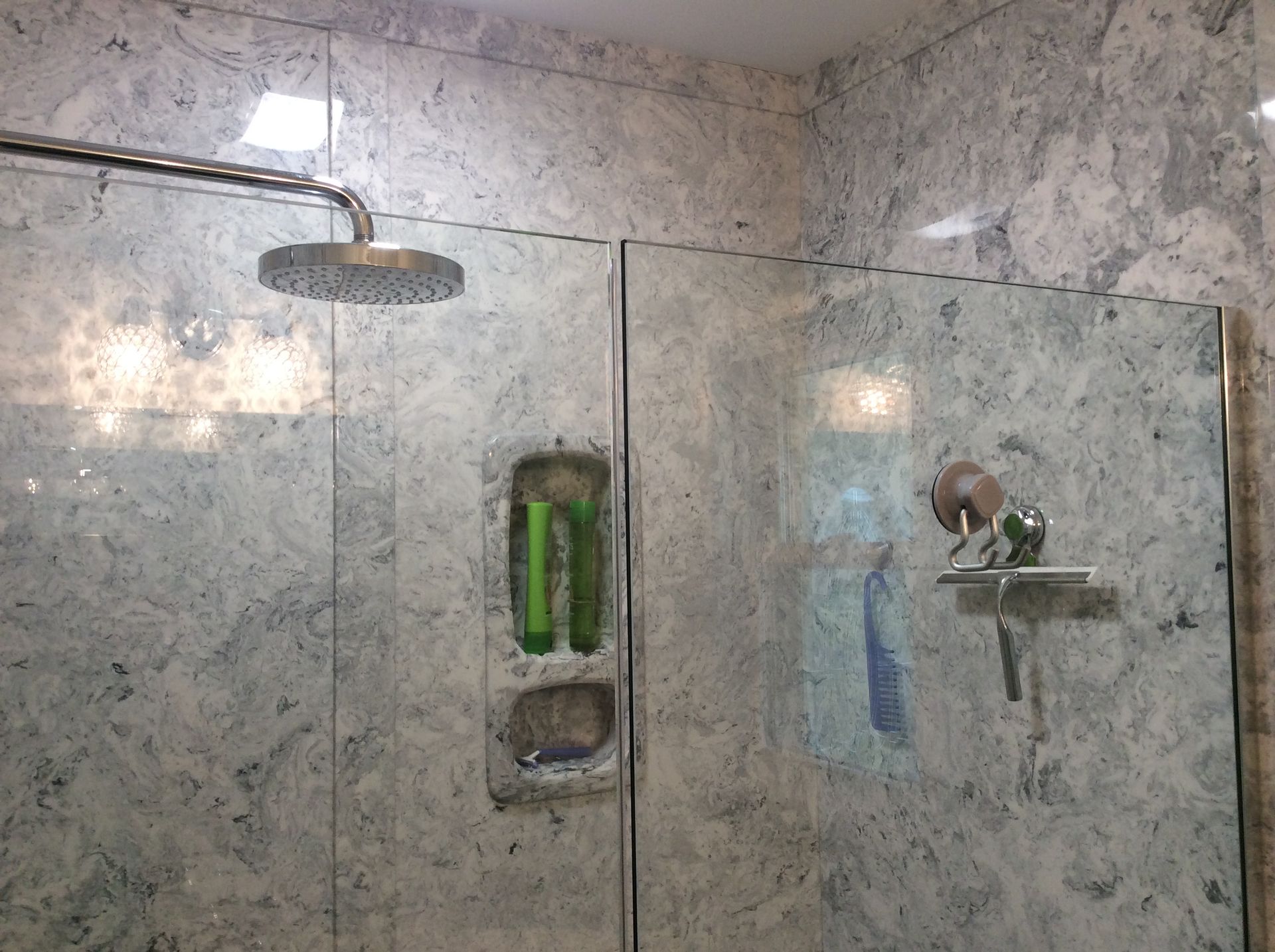 Shower with marble-patterned walls, glass doors, and a round showerhead. Soap and shampoo in a recessed shelf.