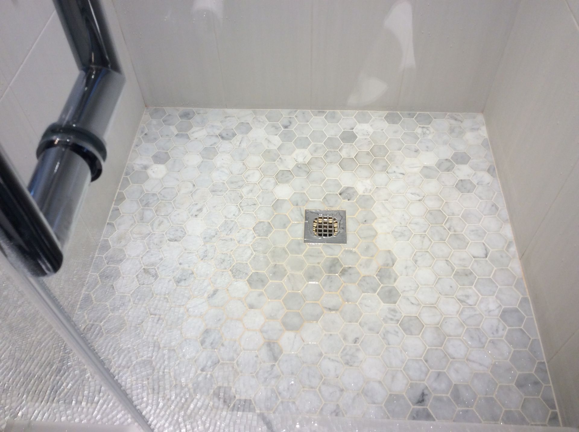 Shower floor with gray hexagon tiles, drain, and chrome plumbing against white walls.