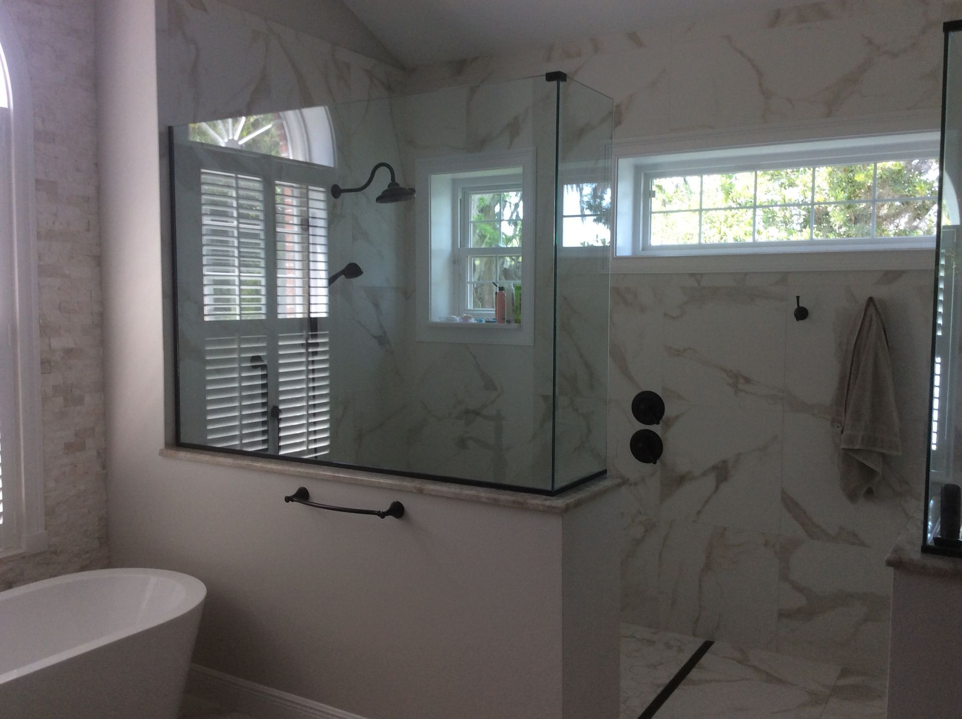 Bathroom with glass shower, marble walls, white tub, and black fixtures. Windows provide natural light.