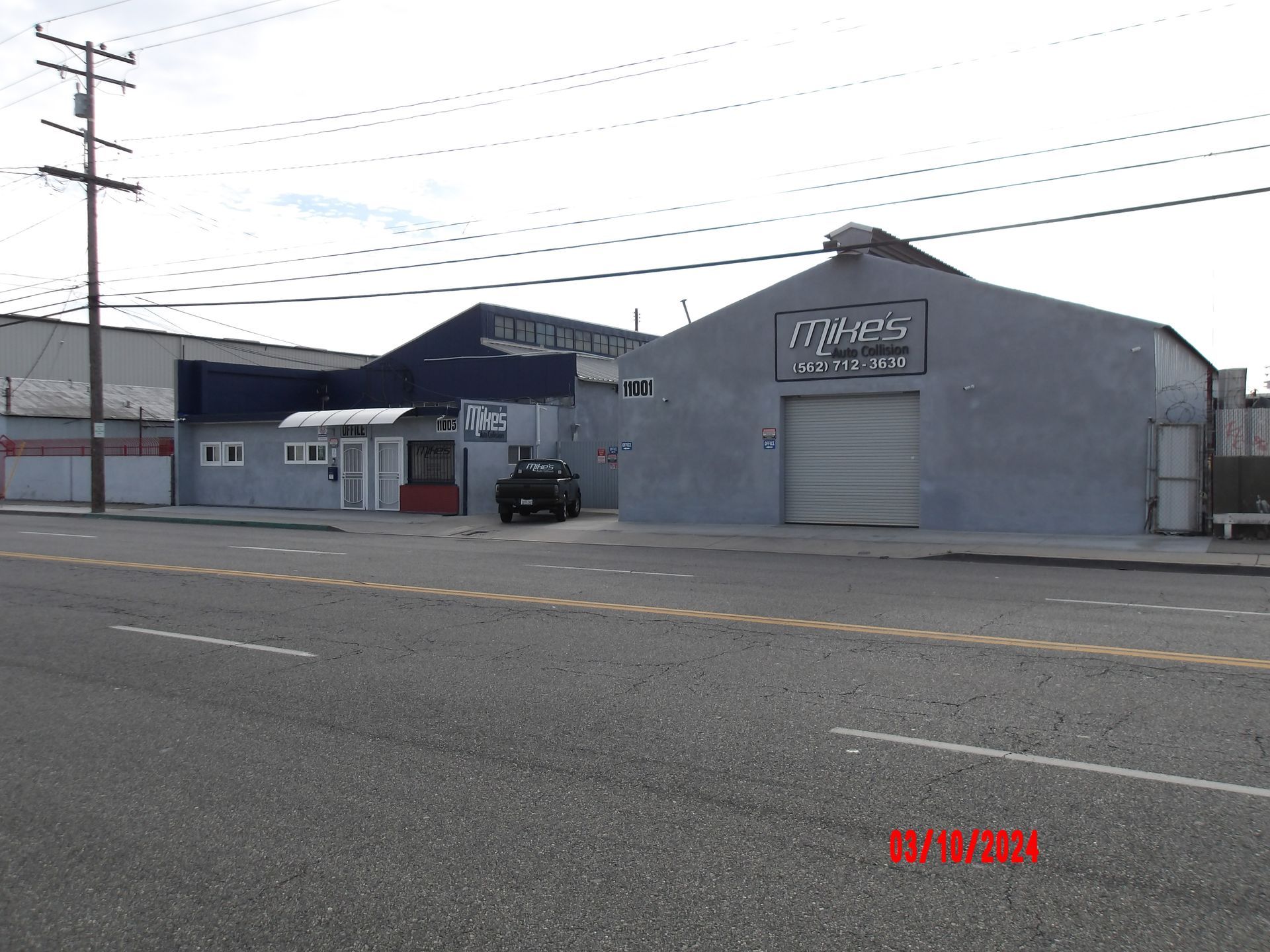 Gray industrial building with roll-up door and small office, parked car, power lines, and street in front.
