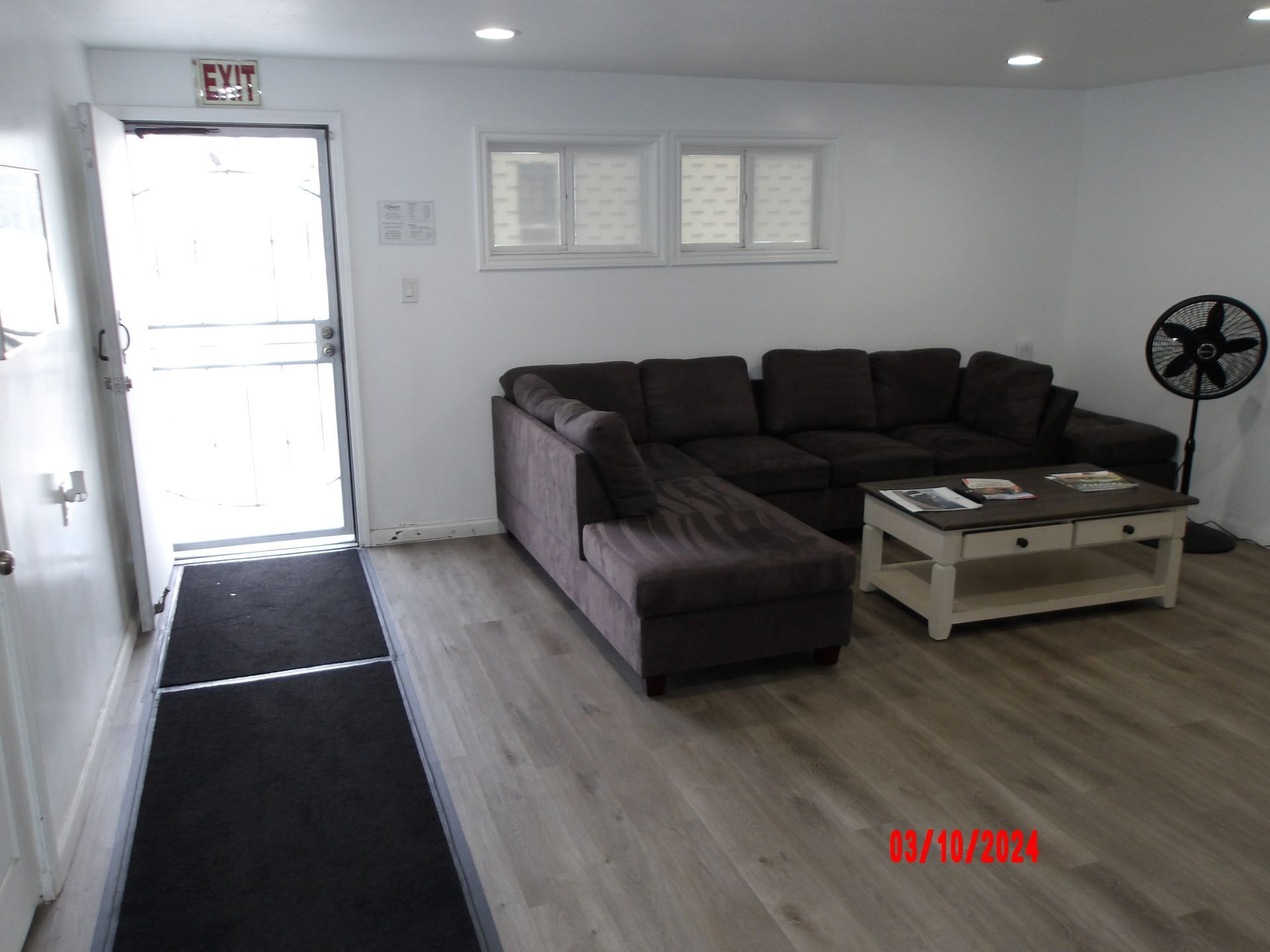 Bright living room with gray sectional, coffee table, fan, and open front door to a black entry rug.