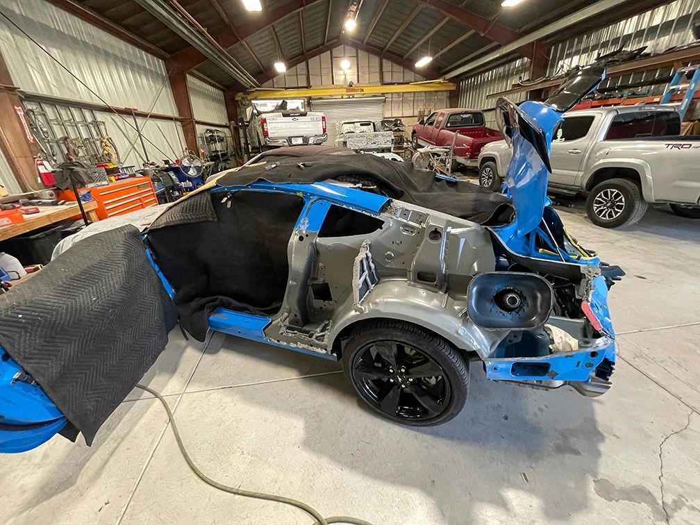 Severely damaged blue sports car in an auto shop, rear and side panels stripped away.