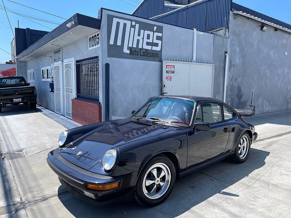 Black Porsche 911 parked outside a gray building with a “Mikes” sign on a sunny street
