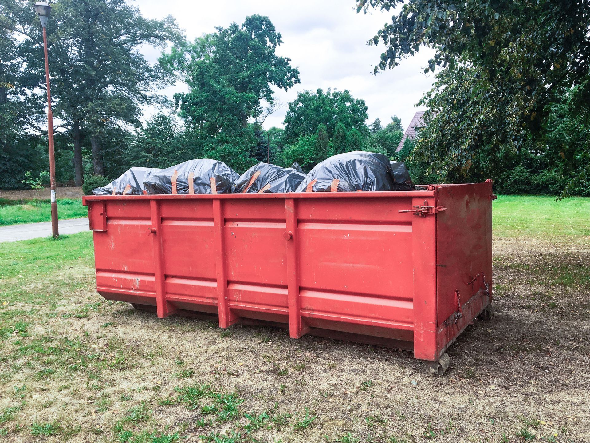Red dumpster filled with black trash bags on grass, trees in background.