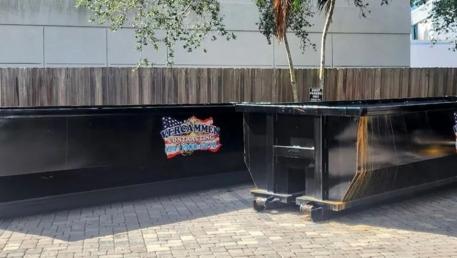 Two black dumpsters with a mural on brick pavers. A wooden fence and tree are in the background.