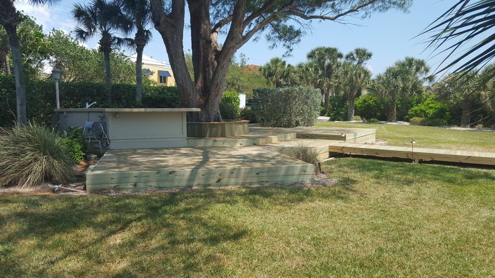A wooden outdoor structure with a raised sandbox area sits under a large tree in a sunlit backyard lawn.