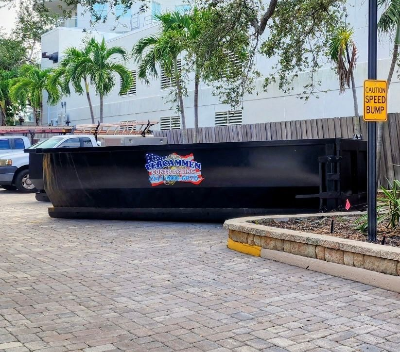A large black dumpster with a patriotic logo sits on a paved lot next to a white pickup truck and a yellow speed bump sign.