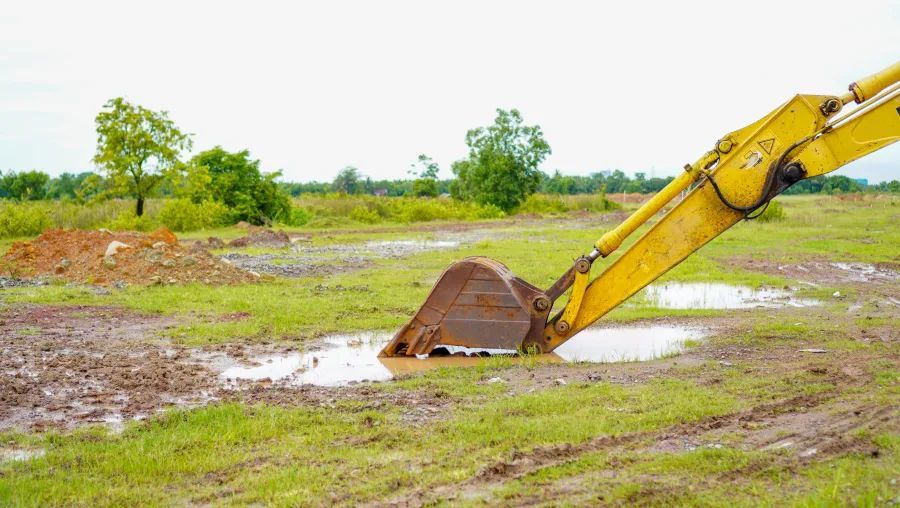 Yellow excavator bucket stuck in mud, working in a grassy, muddy field. Trees in the background.