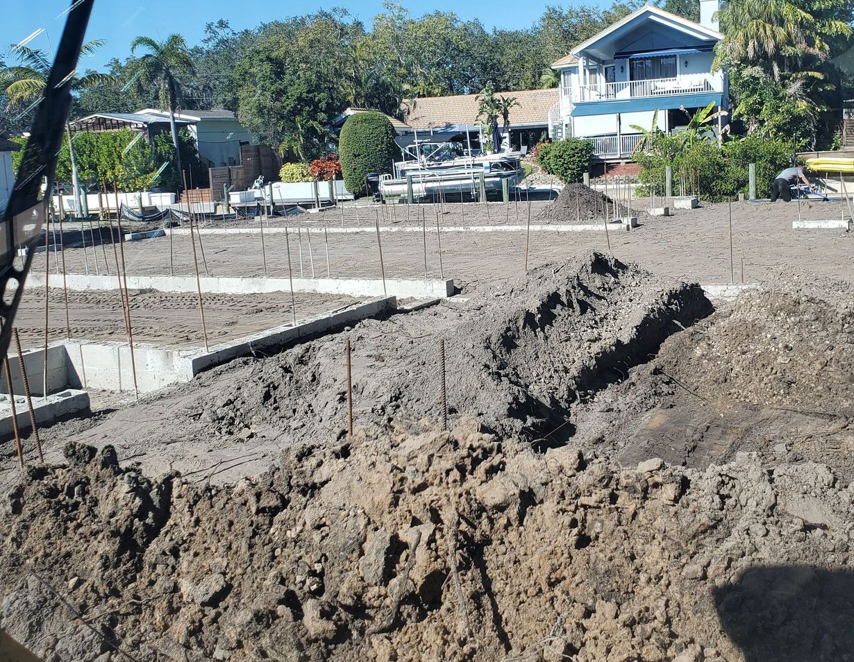 A construction site with concrete foundation walls and trenches in loose soil, with houses and trees in the background.