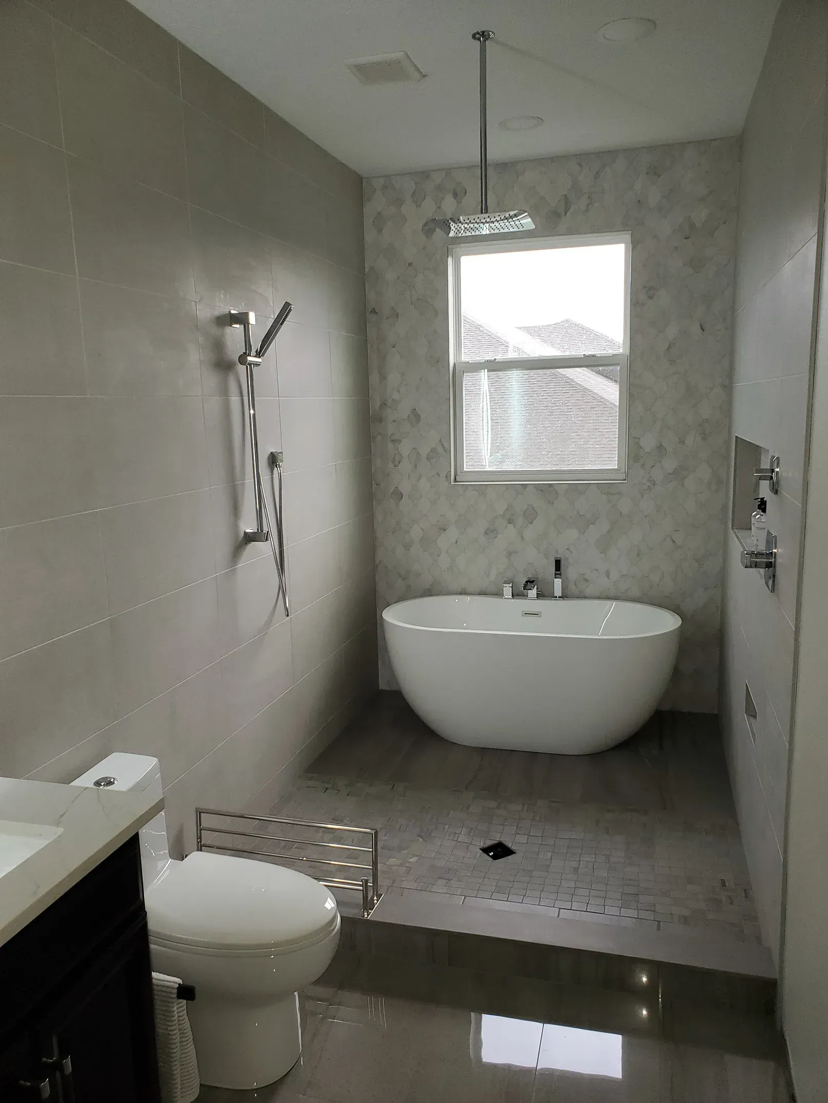 Modern bathroom with a freestanding tub, walk-in shower, toilet, and window. Gray walls and flooring.