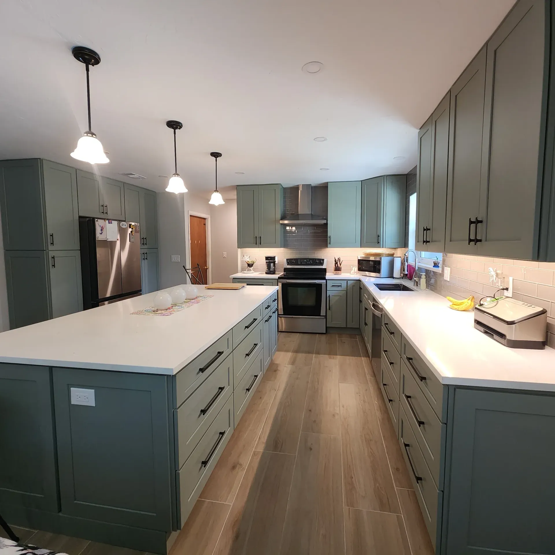 A kitchen with green cabinets, white countertops, and a large island. Wooden floors and recessed lighting are visible.