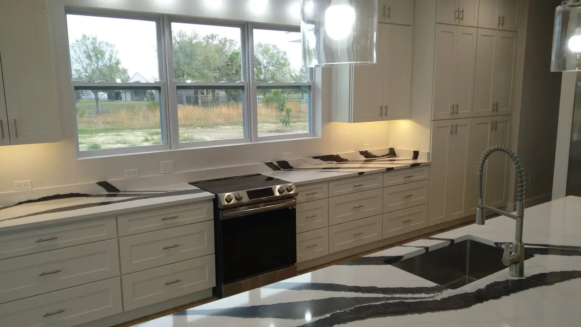 Modern kitchen with white cabinets, marble countertops, stainless steel appliances, and a window overlooking a field.