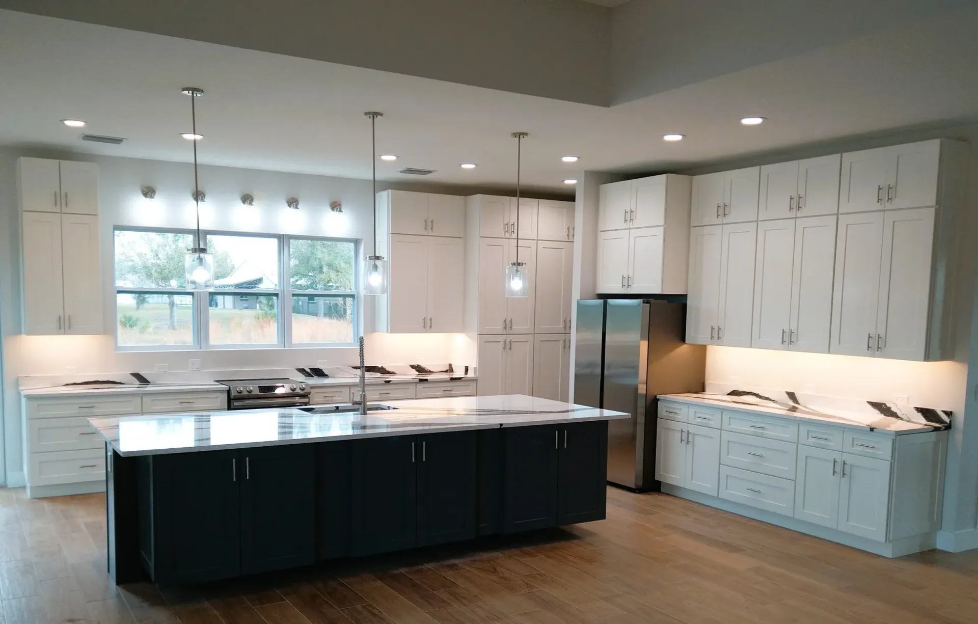 Modern kitchen with white cabinets, dark island, stainless steel refrigerator, and wood flooring.