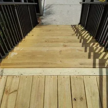 Wooden deck stairs with dark railings, leading downwards to a landing. Sunlight casts shadows.