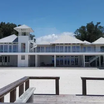 White beach house with tower, spiral staircase, on a sandy beach under a blue sky.