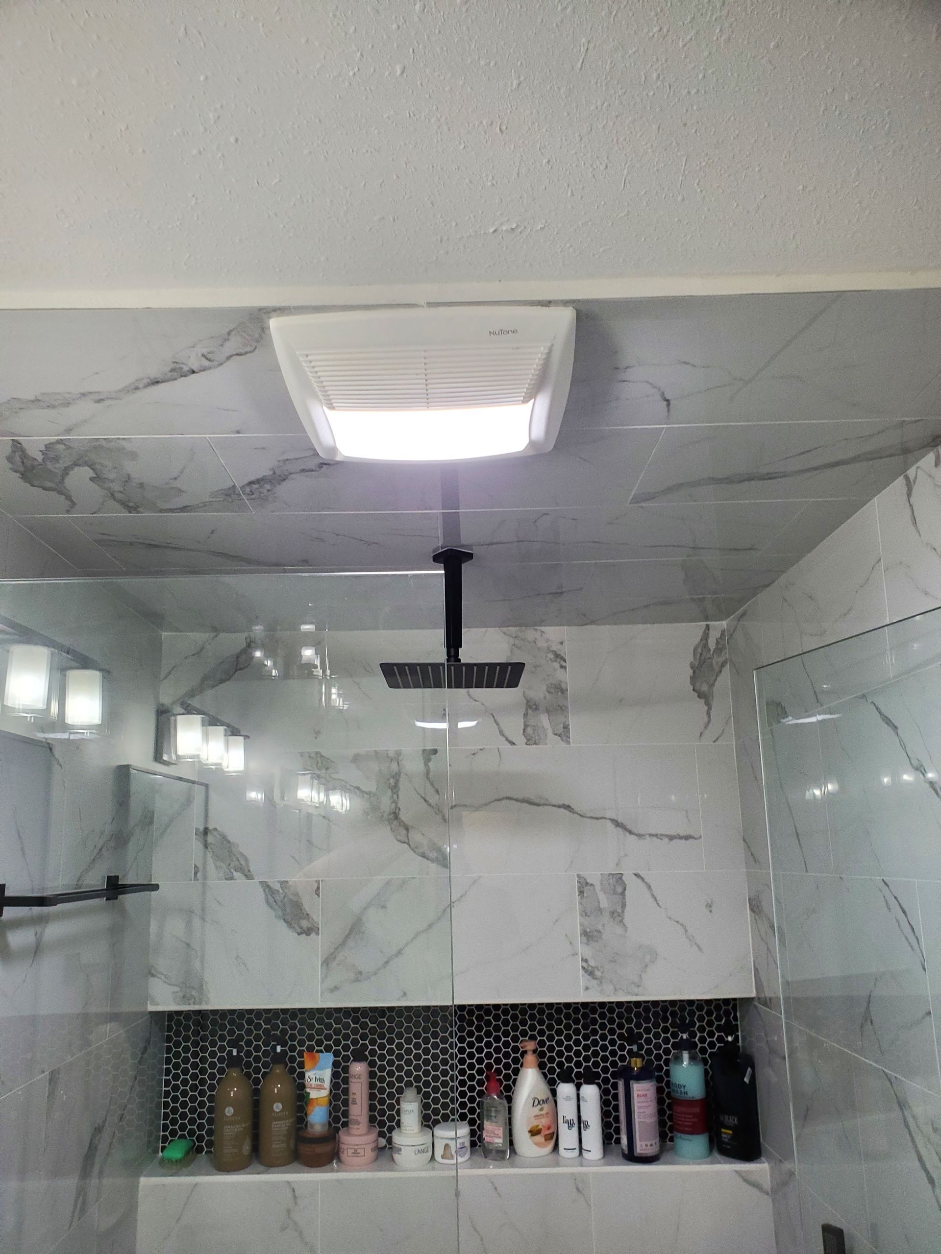 A bathroom shower area with a square ceiling light and a black rainfall showerhead above marble-patterned tiled walls.