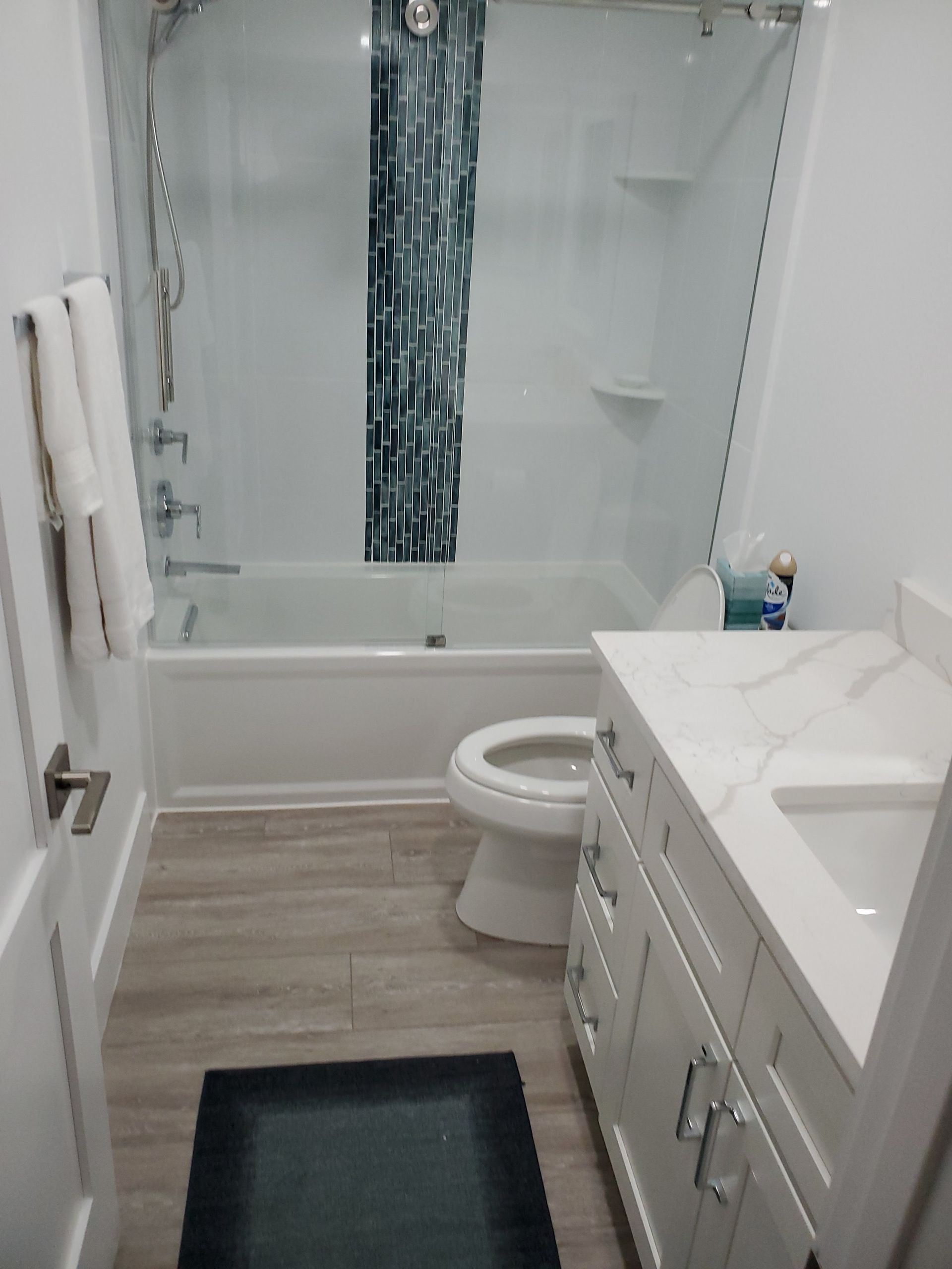 A bathroom with wood-look flooring, a white vanity, a toilet, and a bathtub with a vertical patterned teal shower curtain.