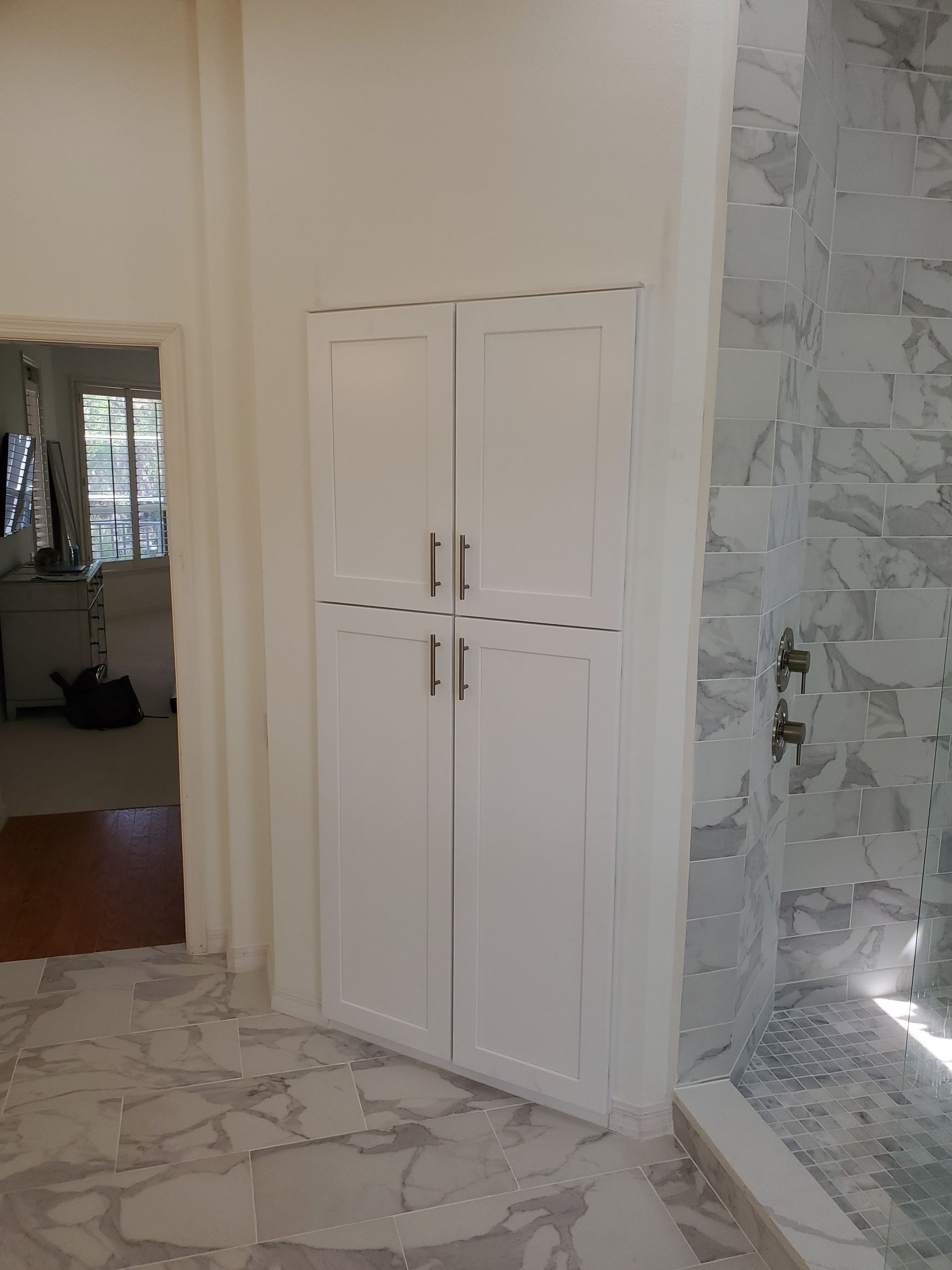 White cabinets with tall doors built into a wall next to a marble shower, with a doorway visible to the left.