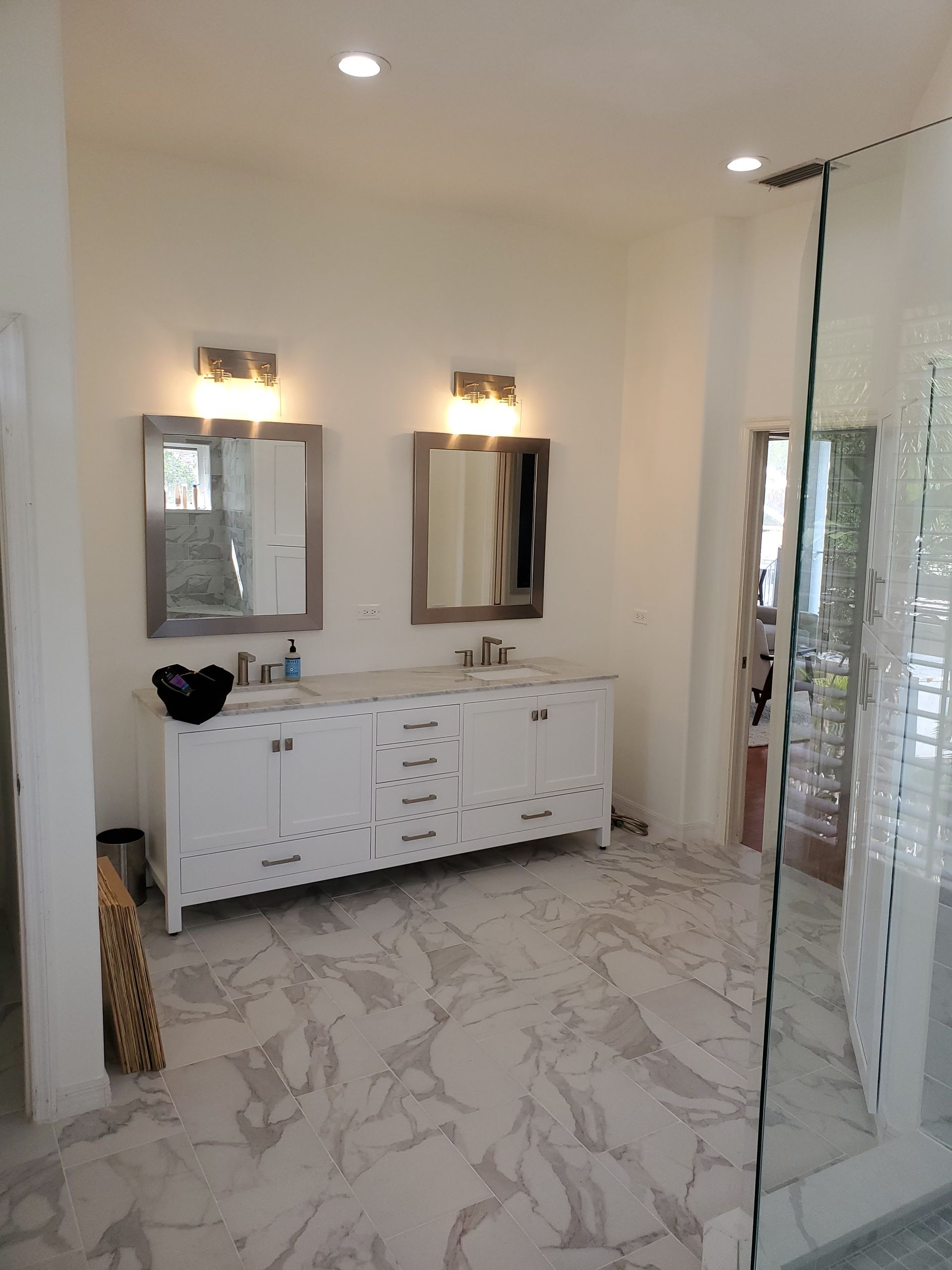 A bright bathroom features a double-sink white vanity, two rectangular mirrors, wall lighting, and marbled tile flooring.