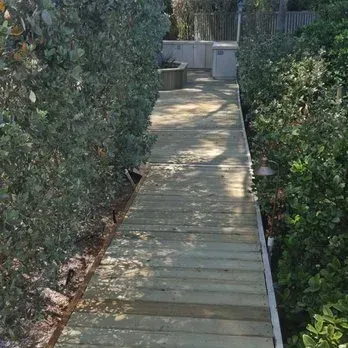 Wooden walkway between tall green hedges, leading toward an outdoor kitchen area.