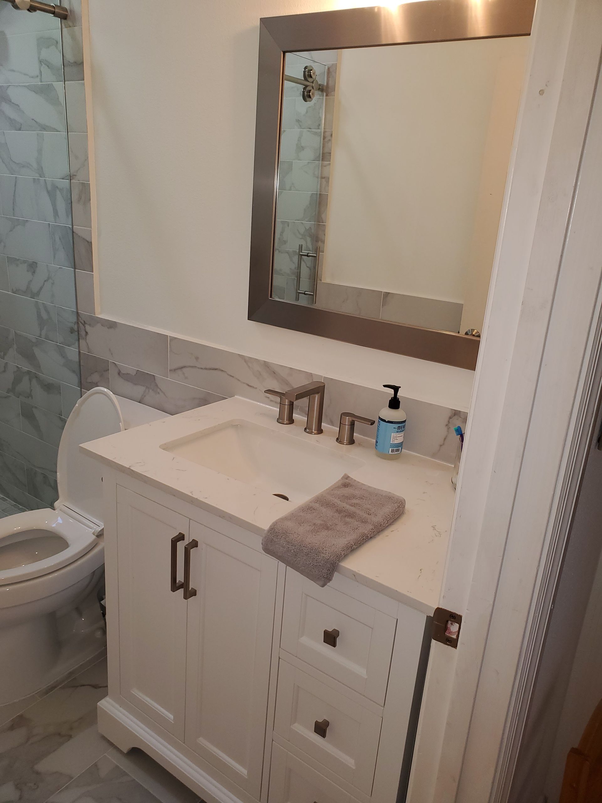 A bathroom vanity with a white cabinet, marble countertop, silver faucet, and mirror, next to a white toilet.