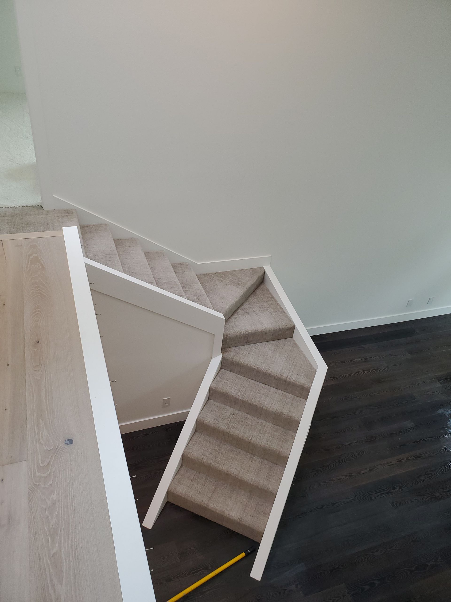 A downward view of beige carpeted stairs with a white banister, transitioning from a light wood floor to a dark floor.