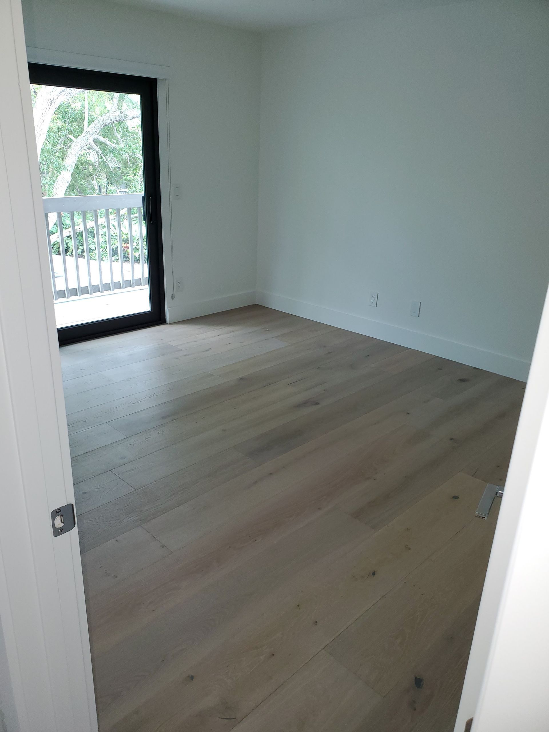 An empty room with light wood floors, white walls, and a sliding glass door opening to a balcony with trees.