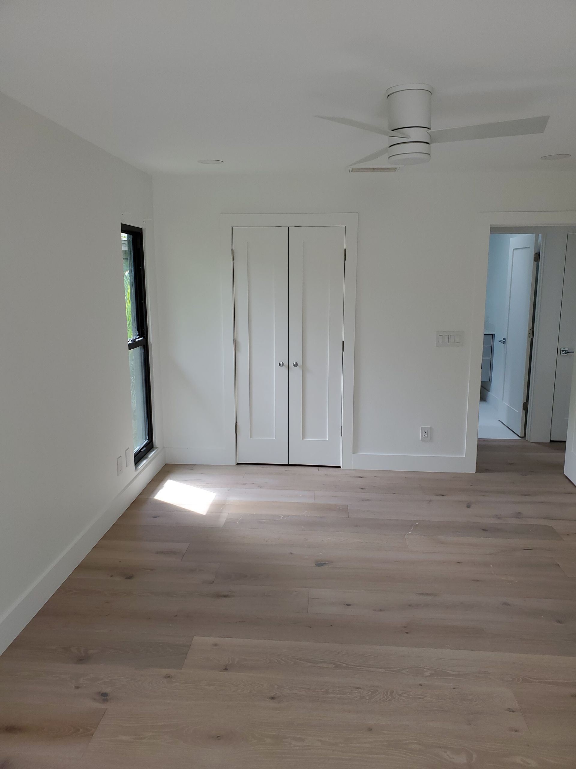 A bright, modern room with white walls, light wood flooring, a ceiling fan, a window, and closed closet doors.