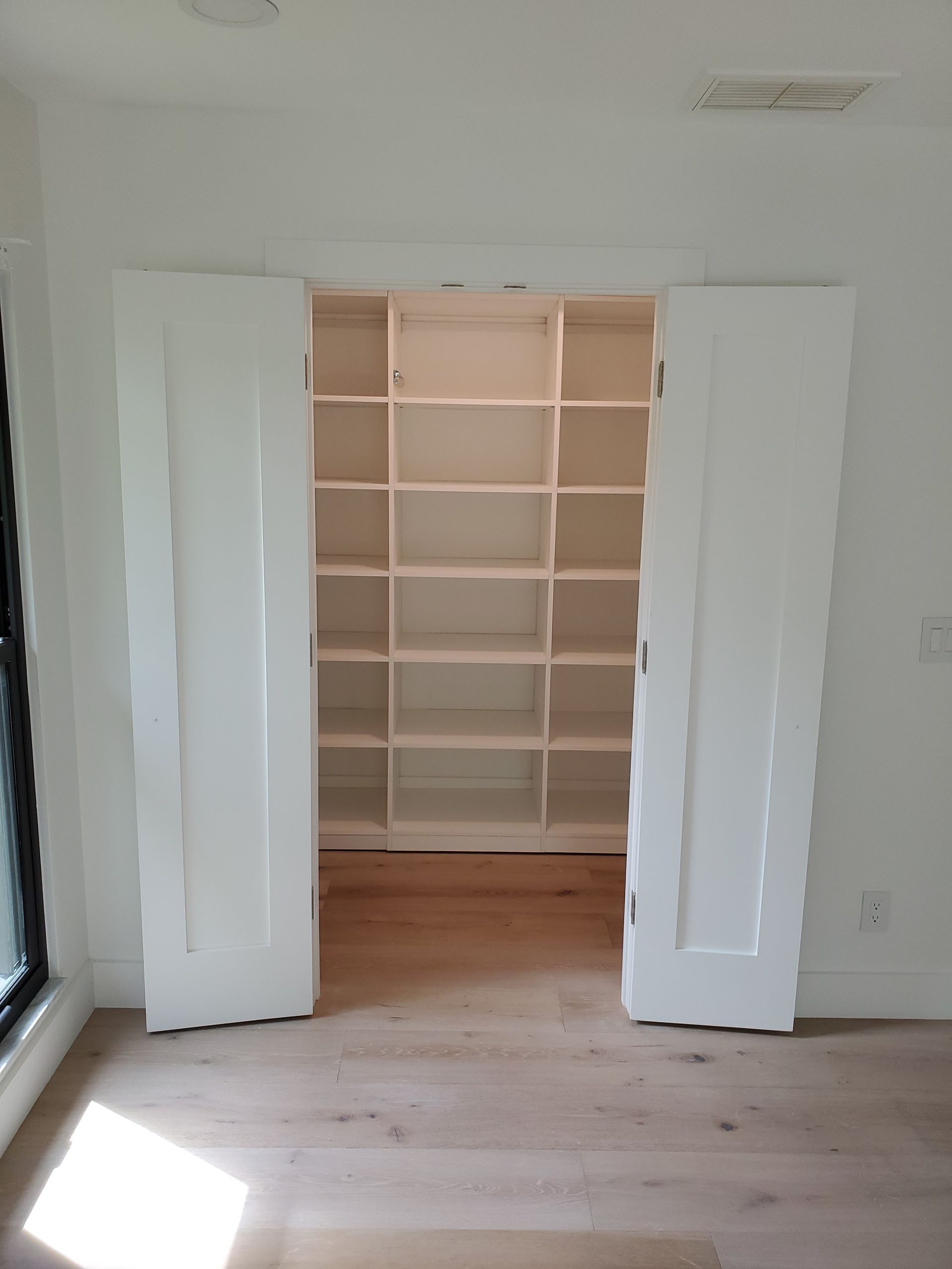 Open white doors reveal a walk-in pantry with white wooden shelving against a back wall, set in a room with wood flooring.