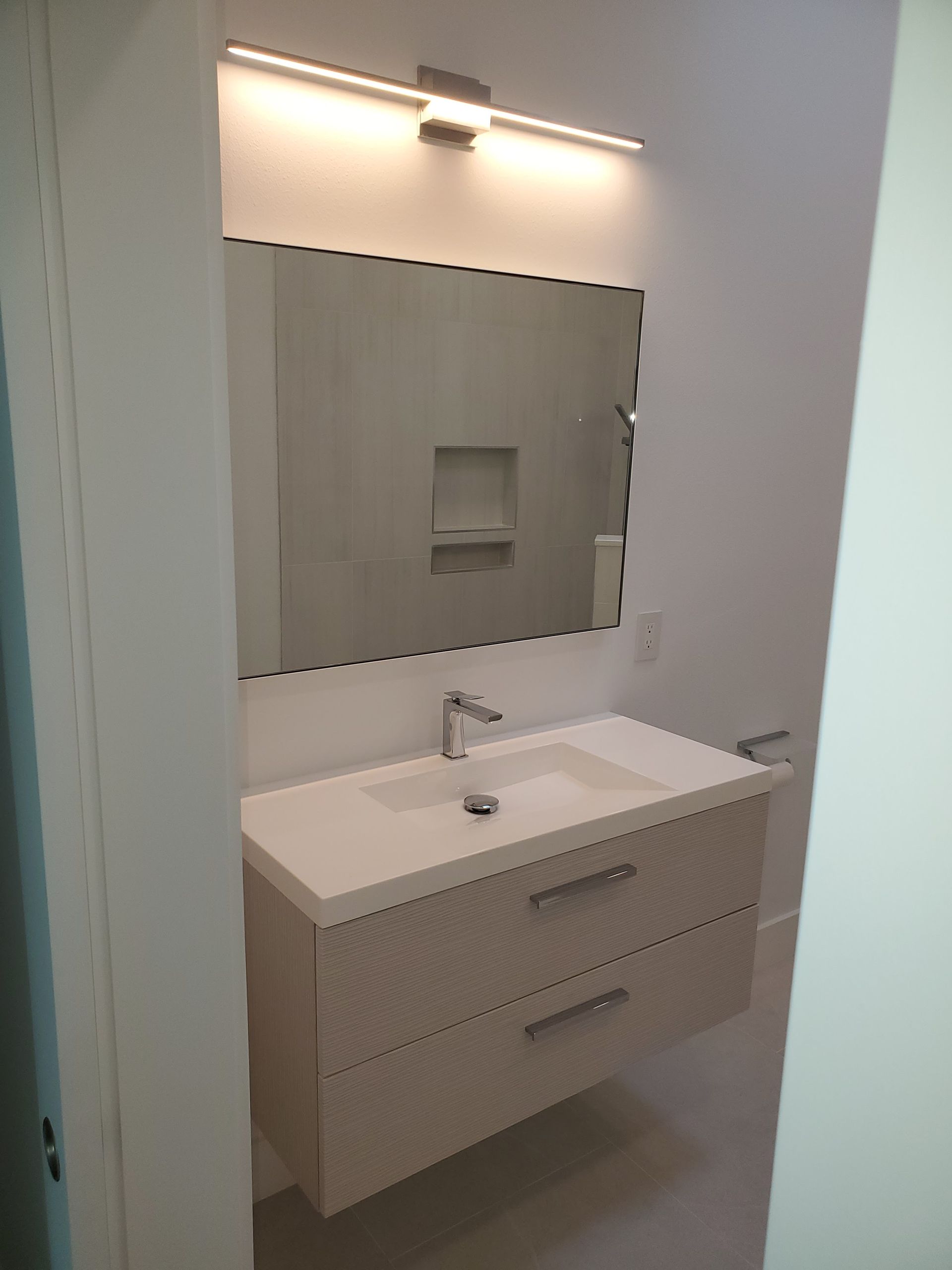A modern bathroom vanity with a white countertop, wall-mounted cabinet, rectangular mirror, and an LED light fixture.