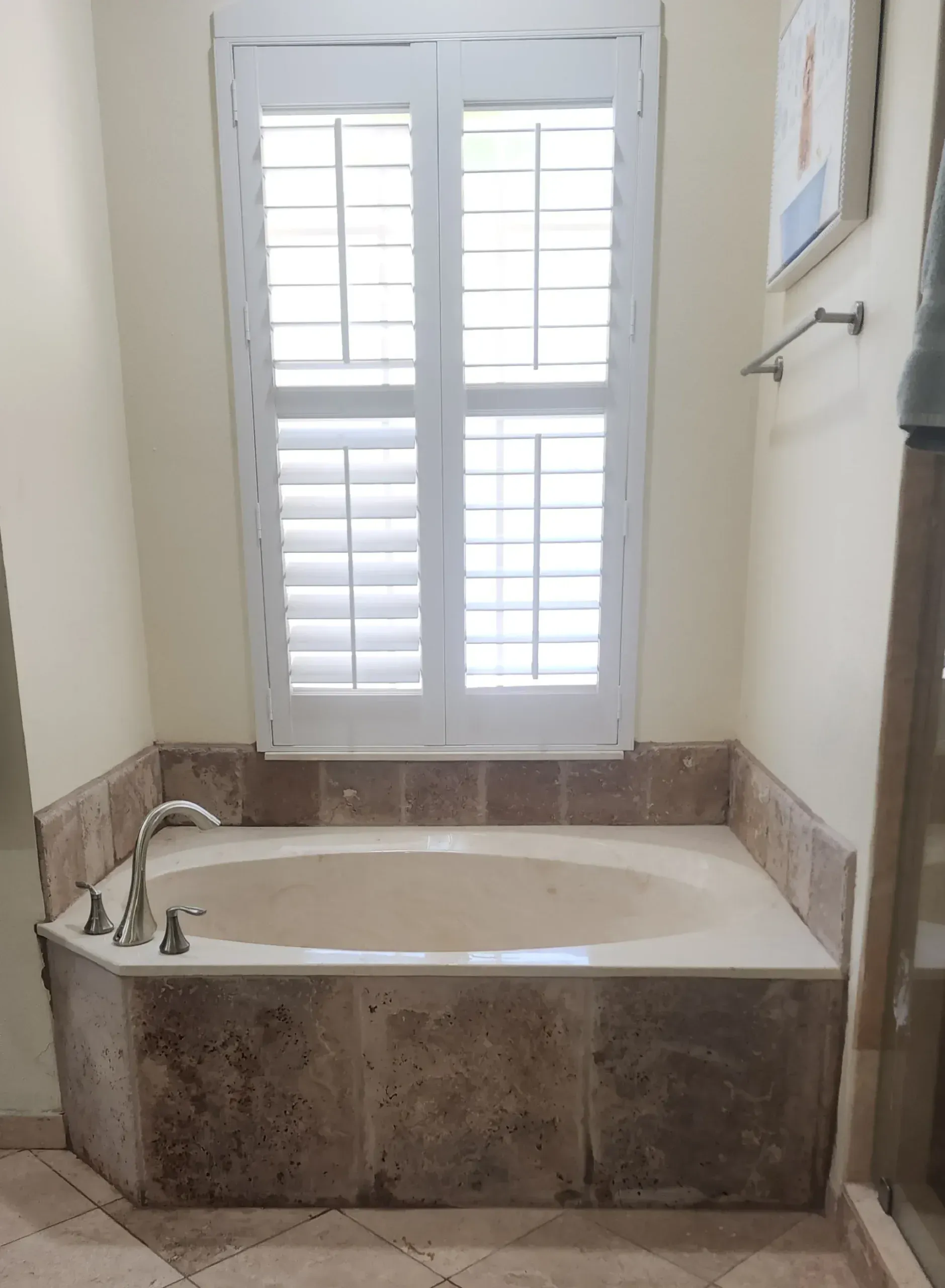 Bathroom with a built-in beige bathtub, marble surround, and shuttered window.