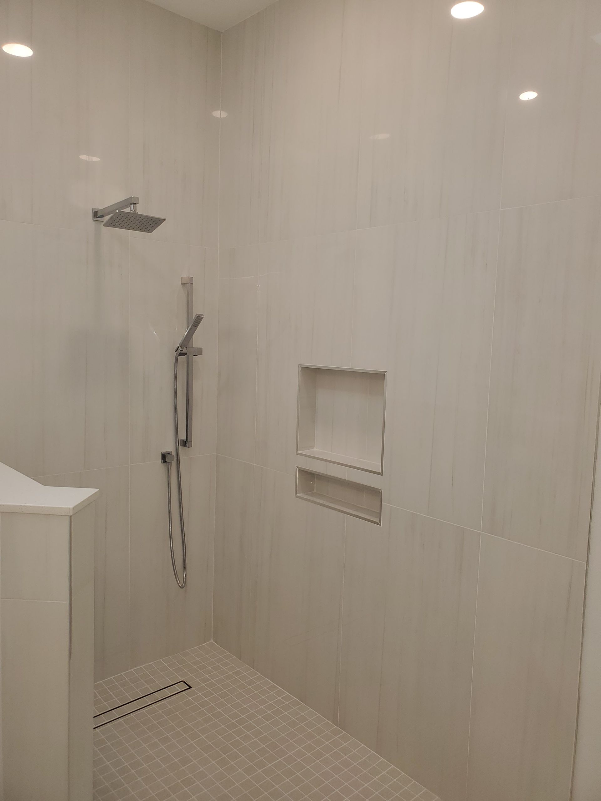 A modern walk-in shower with light gray tiles, a rainfall showerhead, a handheld sprayer, and a built-in storage niche.